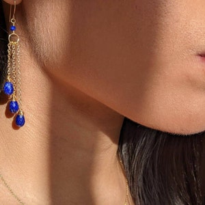 May include: Gold-toned earrings with a cascading design. Each earring features a small gold hoop, delicate chains, and three deep blue teardrop-shaped gemstones. The earrings are suspended from a gold hook.