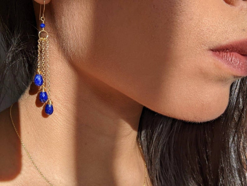 May include: Gold-toned earrings with a cascading design. Each earring features a small gold hoop, delicate chains, and three deep blue teardrop-shaped gemstones. The earrings are suspended from a gold hook.