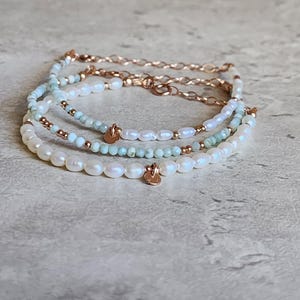 May include: A stack of three beaded bracelets. The bracelets feature white pearls, light blue beads, and gold-colored chain and accents. Each bracelet has a small, gold-colored charm. The bracelets are displayed on a light gray surface.