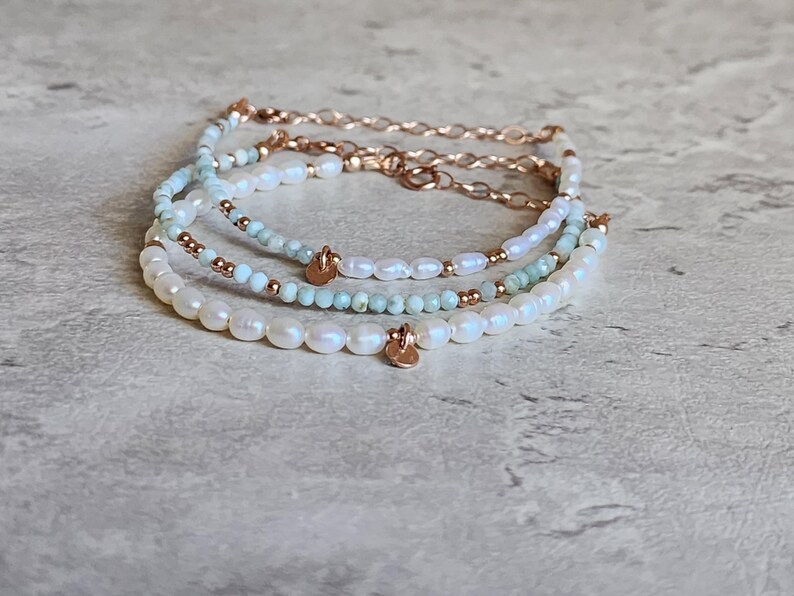 May include: A stack of three beaded bracelets. The bracelets feature white pearls, light blue beads, and gold-colored chain and accents. Each bracelet has a small, gold-colored charm. The bracelets are displayed on a light gray surface.