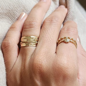 May include: A set of four gold stacking rings on one finger, featuring smooth and twisted bands. On the adjacent finger, a gold ring with a central star-shaped setting and small clear stones along the band, creating a delicate layered appearance.