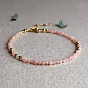 May include: A delicate bracelet made with small pink opal beads and gold accents. The bracelet is strung on a clear elastic cord.