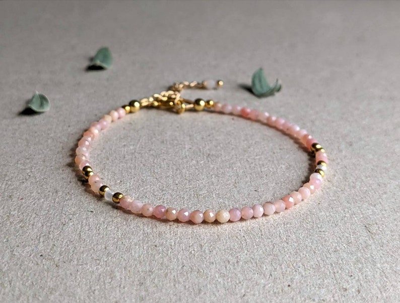 May include: A delicate bracelet made with small pink opal beads and gold accents. The bracelet is strung on a clear elastic cord.