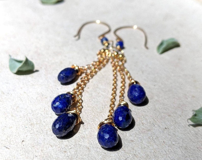 May include: Gold-tone dangle earrings featuring faceted, deep blue lapis lazuli teardrop-shaped beads. The earrings have a delicate chain design and are suspended from curved ear wires. The jewelry is displayed on a neutral surface.