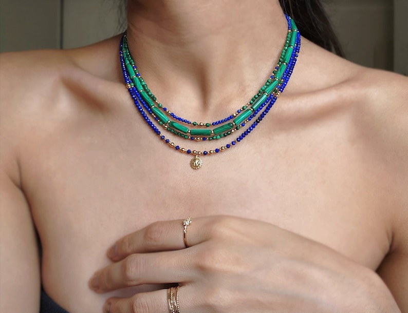 May include: A layered necklace with green and blue beads and a small gold pendant. The necklace is worn by a person with fair skin.