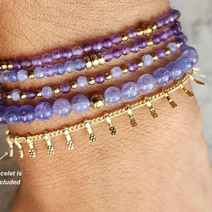 May include: A set of five purple and gold beaded bracelets on a person's wrist. The bracelets are made of amethyst and gold beads. The gold bracelet is not included.