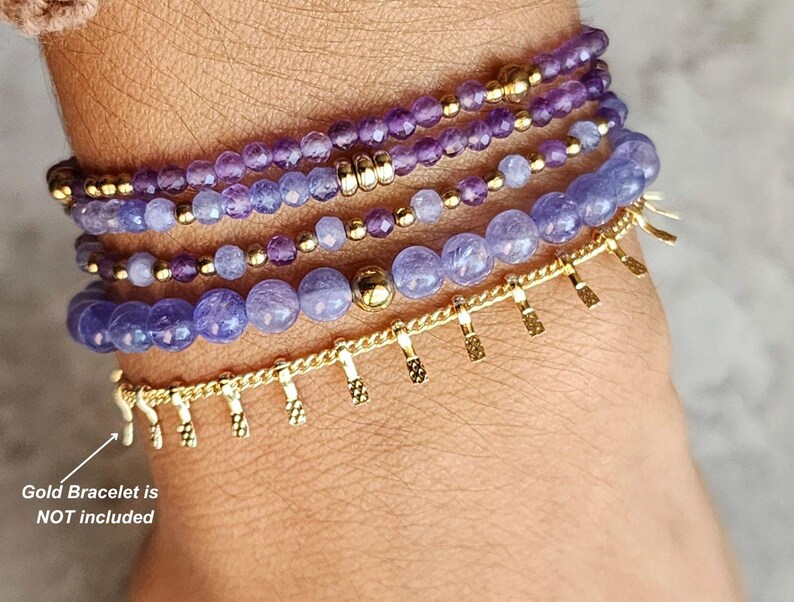 May include: A set of five purple and gold beaded bracelets on a person's wrist. The bracelets are made of amethyst and gold beads. The gold bracelet is not included.