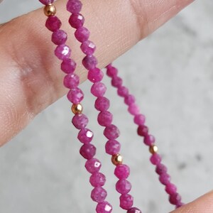 Pu&ograve; includere: Primo piano di una delicata collana con piccole perline sfaccettate color rubino infilate insieme. Perline color oro sono intervallate lungo la collana. La collana &egrave; tenuta su uno sfondo neutro.