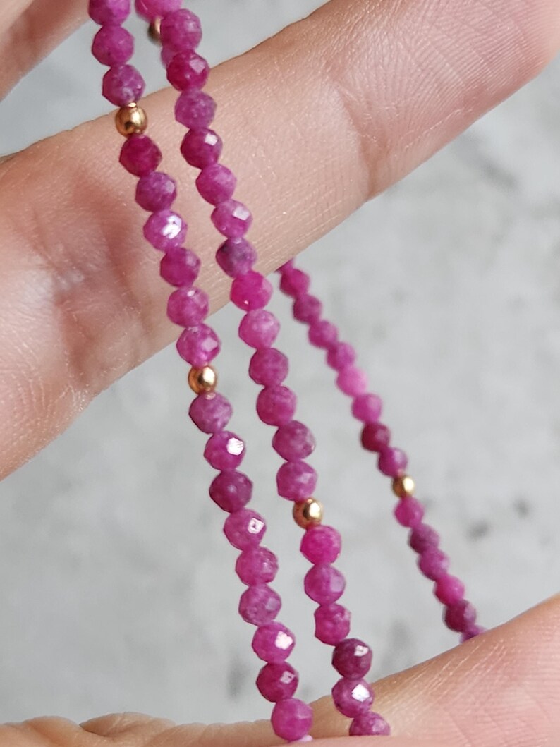 Pu&ograve; includere: Primo piano di una delicata collana con piccole perline sfaccettate color rubino infilate insieme. Perline color oro sono intervallate lungo la collana. La collana &egrave; tenuta su uno sfondo neutro.