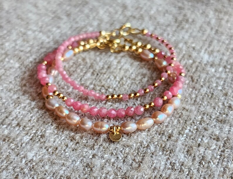 Puede incluir: Tres pulseras apiladas con cuentas rosas y doradas. La pulsera superior tiene peque&ntilde;as cuentas rosas, la del medio tiene cuentas rosas m&aacute;s grandes y la inferior tiene perlas rosas. Todas las pulseras tienen detalles dorados.
