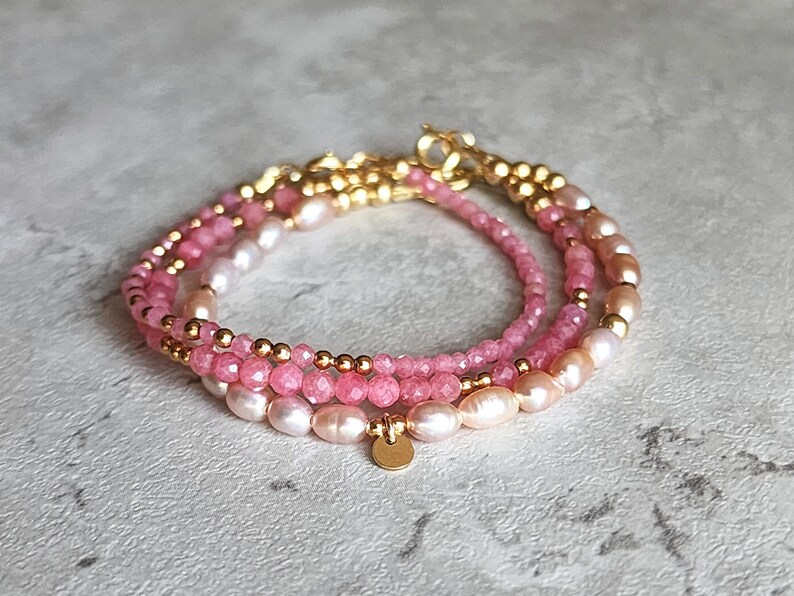 Puede incluir: Pulsera de tres hilos con perlas rosas y melocot&oacute;n, peque&ntilde;as cuentas rosas y detalles dorados. La pulsera tiene un dise&ntilde;o delicado con un peque&ntilde;o dije dorado. La joya se exhibe sobre una superficie gris claro.