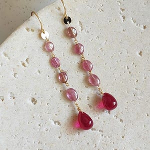 Puede incluir: Pendientes colgantes con ganchos y cadenas doradas. Cada pendiente presenta una serie de cuentas de color rosa y magenta, que culminan en una gema magenta en forma de lágrima. Los pendientes se exhiben sobre una superficie texturizada de color blanquecino.