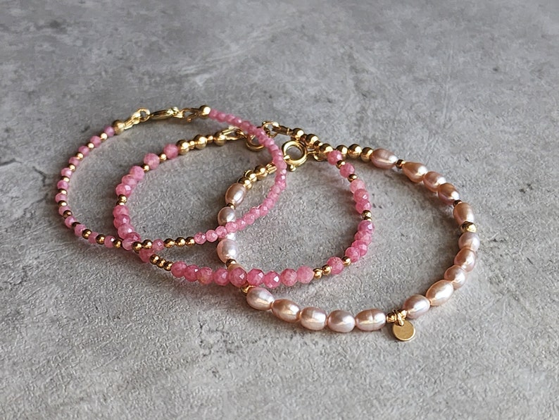 Puede incluir: Tres pulseras delicadas con piedras rosas y perlas. Las pulseras presentan cuentas y cierres dorados. Una pulsera est&aacute; hecha de peque&ntilde;as piedras rosas facetadas, las otras tienen perlas rosas.
