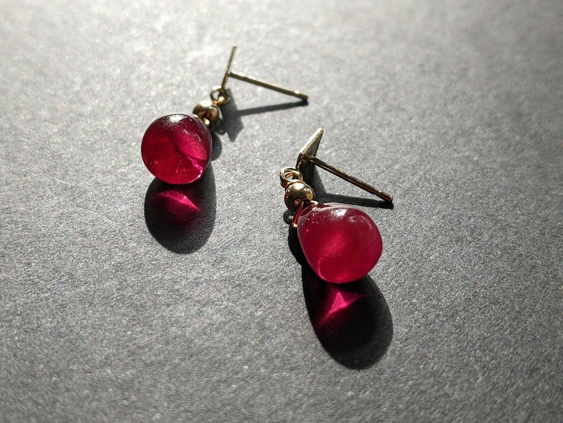 May include: A pair of earrings with red gemstone drops. The earrings have gold posts and are a simple and elegant design.