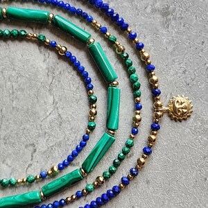 May include: A layered necklace with blue lapis lazuli beads, green malachite beads, and gold beads. The necklace has a gold sun charm.