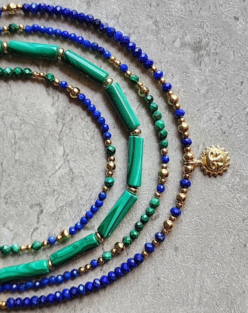 May include: A layered necklace with blue lapis lazuli beads, green malachite beads, and gold beads. The necklace has a gold sun charm.