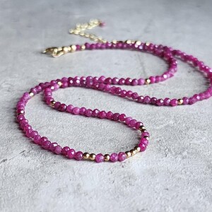 Pu&ograve; includere: Una delicata collana con piccole perline sfaccettate color magenta infilate insieme. Perline color oro sono intervallate lungo la collana, e anche la chiusura e la catena sono dorate. La collana &egrave; esposta su una superficie grigio chiaro.