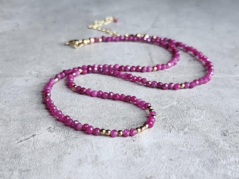 Pu&ograve; includere: Una delicata collana con piccole perline sfaccettate color magenta infilate insieme. Perline color oro sono intervallate lungo la collana, e anche la chiusura e la catena sono dorate. La collana &egrave; esposta su una superficie grigio chiaro.