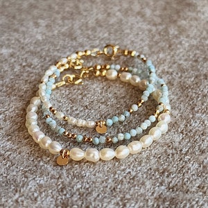 May include: Three layered bracelets with pearls, small gold beads, and light blue beads. Each bracelet has a gold clasp and a small gold disc charm. The bracelets are stacked on a neutral-colored surface.