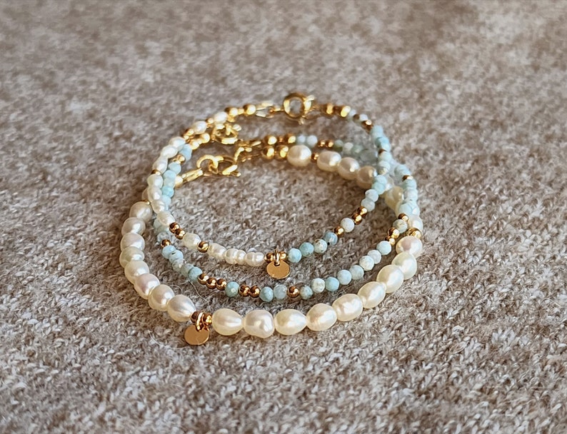 May include: Three layered bracelets with pearls, small gold beads, and light blue beads. Each bracelet has a gold clasp and a small gold disc charm. The bracelets are stacked on a neutral-colored surface.