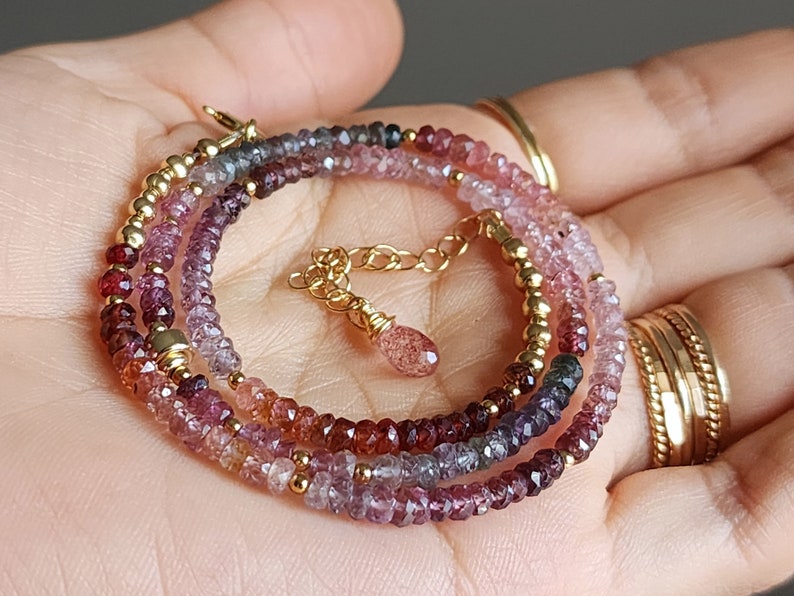 May include: A multi-strand necklace featuring faceted gemstones in pink, purple, and red hues, with small gold beads. The necklace has a gold clasp and a small charm. The jewellery is displayed on a hand.