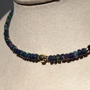 Op de afbeelding: Een ketting met kleine, gefacetteerde donkerblauwe en zwarte opaal kralen met iriserende groene en rode flitsen. De streng is geaccentueerd met een enkele gepolijste goudkleurige cilindrische kraal die centraal tussen de donkere kralen zit.