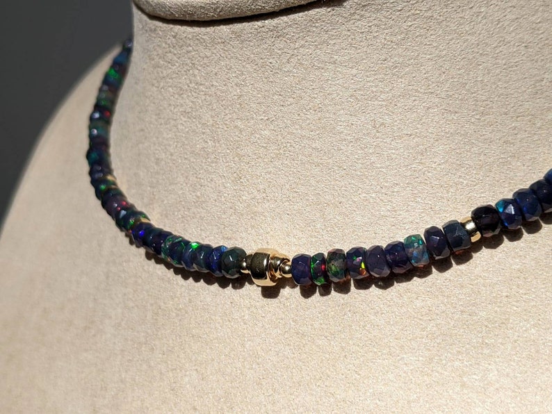 Op de afbeelding: Een ketting met kleine, gefacetteerde donkerblauwe en zwarte opaal kralen met iriserende groene en rode flitsen. De streng is geaccentueerd met een enkele gepolijste goudkleurige cilindrische kraal die centraal tussen de donkere kralen zit.