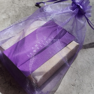 May include: A purple organza gift bag with a drawstring closure holds a rectangular box. The box is wrapped in purple paper with the text "AMTID JEWELLERY" printed in white. The bag and box are on a gray surface.
