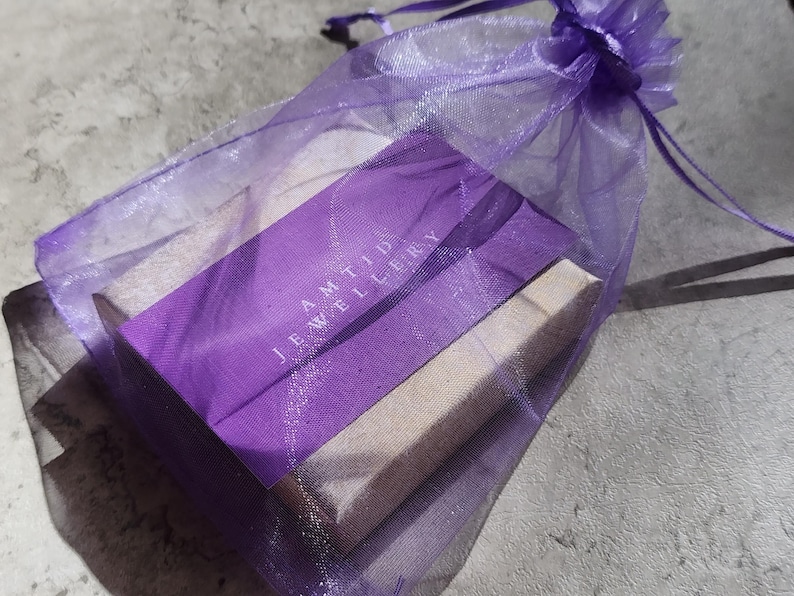 May include: A purple organza gift bag with a drawstring closure holds a rectangular box. The box is wrapped in purple paper with the text "AMTID JEWELLERY" printed in white. The bag and box are on a gray surface.