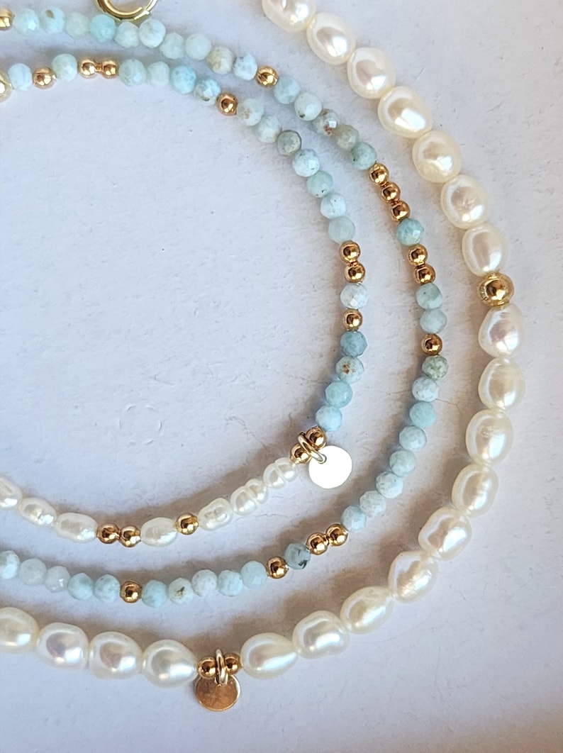May include: Three layered necklaces with pearls, light blue beads, and gold-colored accents. Each necklace has a different design, with a mix of round and irregular-shaped beads. The necklaces are on a white background.