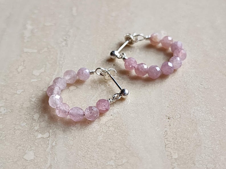 May include: A pair of silver hoop earrings with pink gemstone beads. The earrings are made with small, faceted beads that are strung together to form a circle.