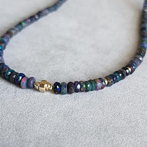 Op de afbeelding: Een ketting bestaande uit een streng kleine, gefacetteerde donkerblauwe en zwarte opaal kralen met iriserende kleurflitsen. Een enkele, gepolijste goudkleurige cilindrische kraal met een centrale rand fungeert als middelpunt tussen de stenen.