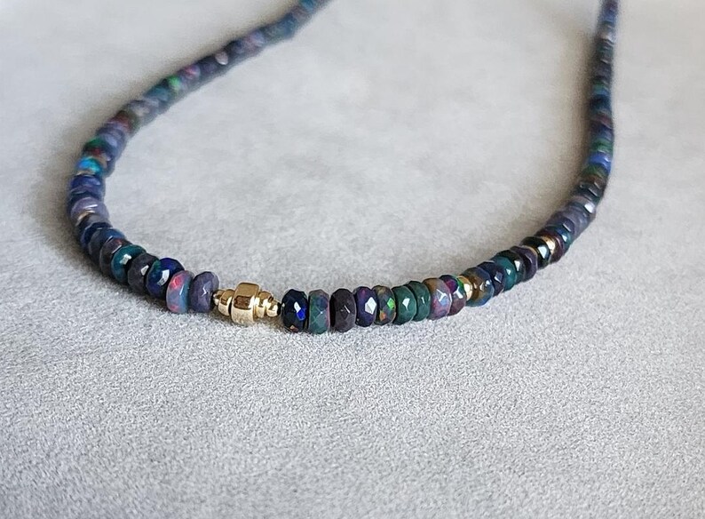 Op de afbeelding: Een ketting bestaande uit een streng kleine, gefacetteerde donkerblauwe en zwarte opaal kralen met iriserende kleurflitsen. Een enkele, gepolijste goudkleurige cilindrische kraal met een centrale rand fungeert als middelpunt tussen de stenen.