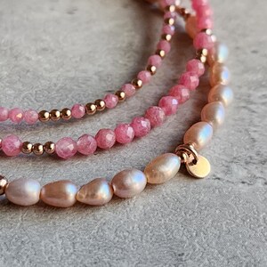 Puede incluir: Primer plano de tres pulseras delicadas. La pulsera superior presenta peque&ntilde;as cuentas rosas, la del medio tiene cuentas rosas facetadas m&aacute;s grandes y la inferior est&aacute; hecha de perlas blanco-rosadas. Todas est&aacute;n acentuadas con cuentas y cierres dorados.