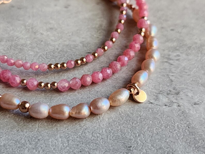 Puede incluir: Primer plano de tres pulseras delicadas. La pulsera superior presenta peque&ntilde;as cuentas rosas, la del medio tiene cuentas rosas facetadas m&aacute;s grandes y la inferior est&aacute; hecha de perlas blanco-rosadas. Todas est&aacute;n acentuadas con cuentas y cierres dorados.