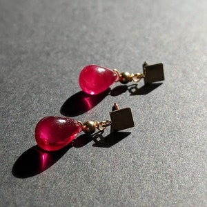 May include: A pair of dangle earrings with teardrop-shaped red gemstone drops and gold square accents.