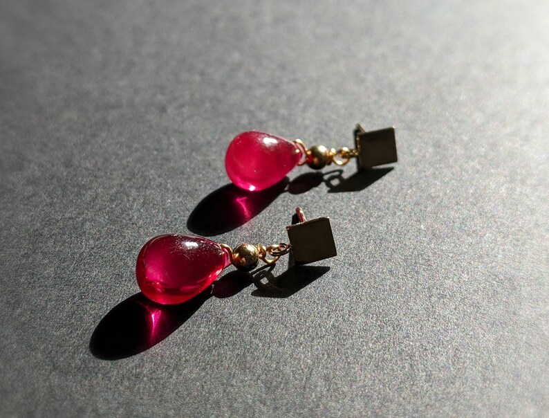 May include: A pair of dangle earrings with teardrop-shaped red gemstone drops and gold square accents.