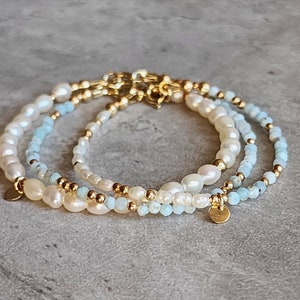 May include: Three delicate bracelets featuring freshwater pearls, light blue beads, and gold-colored accents. Each bracelet has a different design, with a mix of pearl sizes and bead arrangements. The bracelets are displayed on a gray surface.