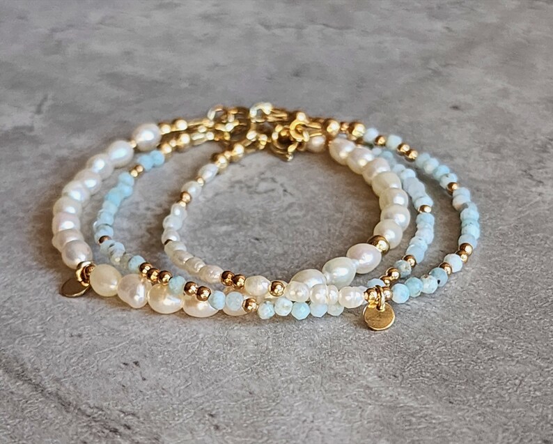 May include: Three delicate bracelets featuring freshwater pearls, light blue beads, and gold-colored accents. Each bracelet has a different design, with a mix of pearl sizes and bead arrangements. The bracelets are displayed on a gray surface.