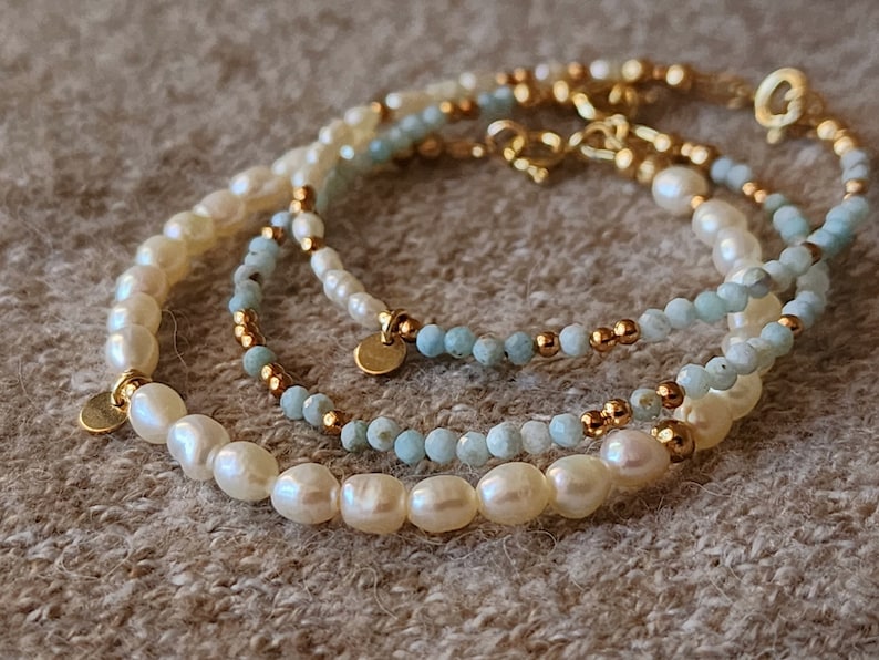 May include: Three delicate bracelets with pearls, light blue beads, and gold accents. The bracelets are layered, creating a stylish and elegant look. Each bracelet has a different design, with varying bead sizes and arrangements. The bracelets are on a neutral background.