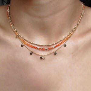 May include: A layered necklace featuring three strands. The top two strands are beaded with small gold and orange beads. The bottom strand has small gold disc charms. The necklace is worn around the neck.