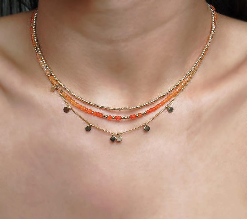 May include: A layered necklace featuring three strands. The top two strands are beaded with small gold and orange beads. The bottom strand has small gold disc charms. The necklace is worn around the neck.