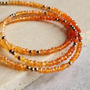 May include: Three strands of orange gemstone bead bracelets with gold-colored accents. The beads are small and faceted, creating a sparkling effect. The bracelets are displayed on a light-colored surface.