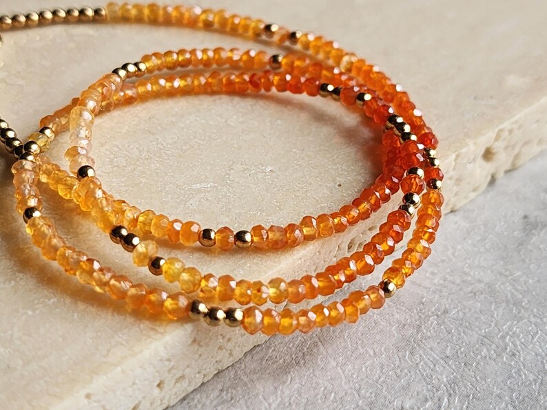 May include: Three strands of orange gemstone bead bracelets with gold-colored accents. The beads are small and faceted, creating a sparkling effect. The bracelets are displayed on a light-colored surface.