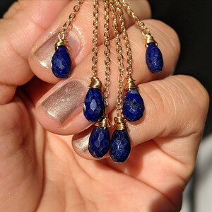 May include: Gold-colored chain earrings with multiple strands, each adorned with a faceted, deep blue teardrop-shaped gemstone. The gemstones are wire-wrapped to the chains. The earrings are displayed against a neutral background.