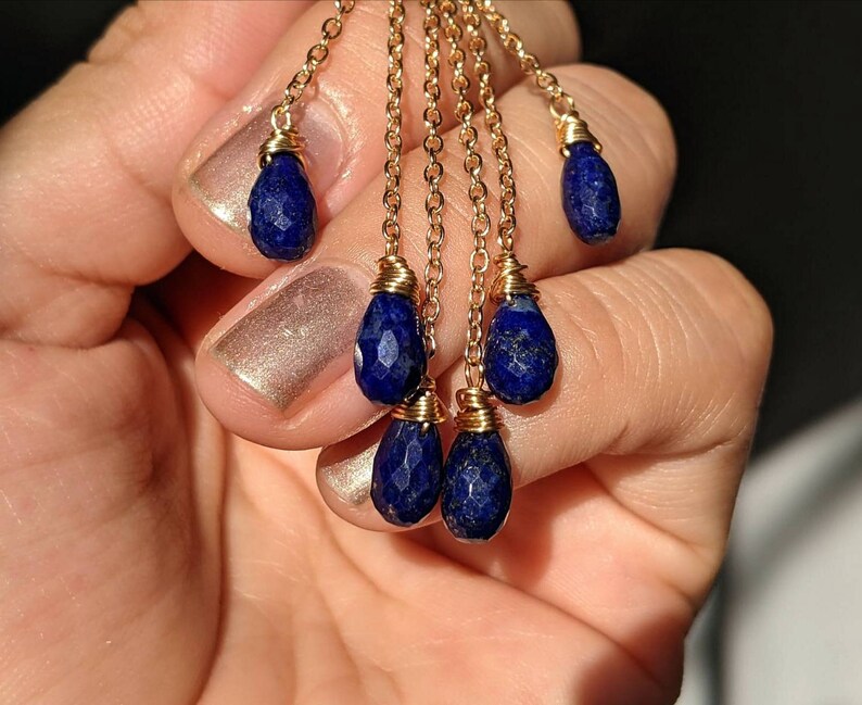 May include: Gold-colored chain earrings with multiple strands, each adorned with a faceted, deep blue teardrop-shaped gemstone. The gemstones are wire-wrapped to the chains. The earrings are displayed against a neutral background.
