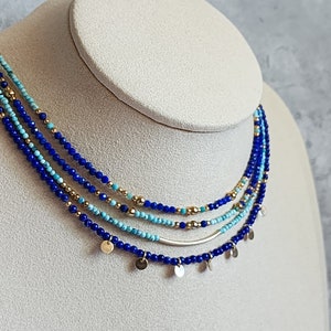 May include: Layered necklace with blue and turquoise beads and gold accents. The necklace features a silver bar and small gold discs hanging from the bottom.