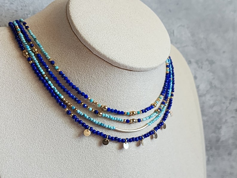May include: Layered necklace with blue and turquoise beads and gold accents. The necklace features a silver bar and small gold discs hanging from the bottom.