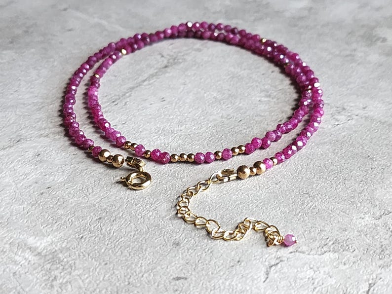 Pu&ograve; includere: Una delicata collana con piccole pietre preziose rosa intenso sfaccettate. Perline color oro e una chiusura e catena di estensione color oro aggiungono contrasto. La collana &egrave; esposta su una superficie strutturata grigio chiaro.