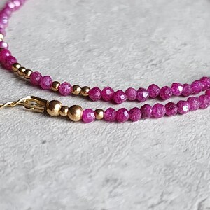 Pu&ograve; includere: Primo piano di una delicata collana con piccole pietre preziose rosa intenso sfaccettate e perline color oro. La collana ha una chiusura e una catena color oro. Il gioiello &egrave; posto su uno sfondo strutturato bianco sporco.